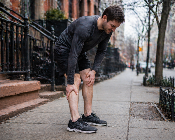 Male runner stopped on a New York City sidewalk, bent over and clutching his knee and shin in pain, depicting a running or walking injury in an urban setting.